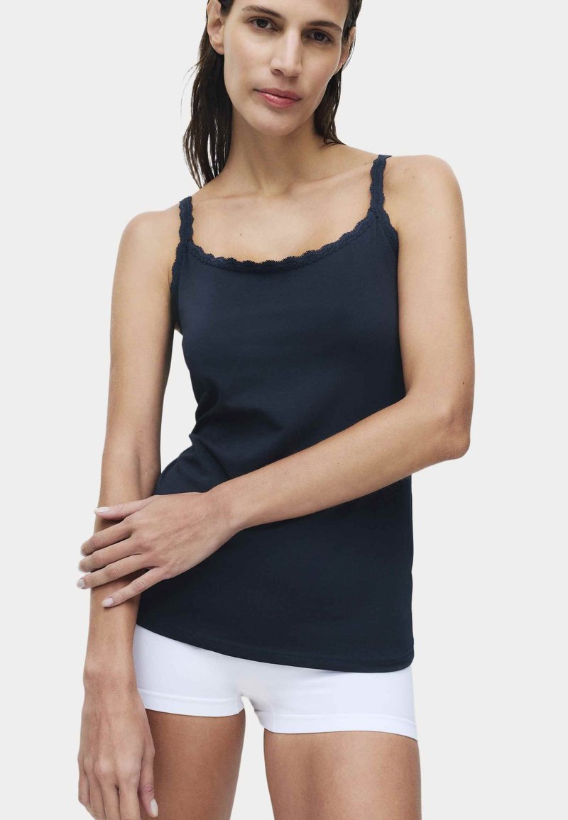 Jeune femme aux cheveux foncés portant un caraco noir orné de dentelle et un short blanc, posant les bras croisés devant un fond uni.