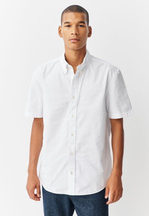 Vīrietis stāv pret vienkāršu fonu, ģērbies baltā īsroku pogātā kreklā un tumši zilās džinsās, skatās taisni priekšā.