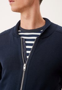 Granatowa dzianinowa kurtka z żebrowanym kołnierzem, wyposażona w srebrny zamek błyskawiczny i teksturowany panel, zestawiona z pasiastą koszulą pod spodem.