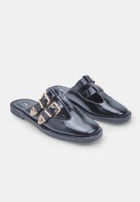 Pair of black shiny slip-on shoes with double gold buckles and cutout details, flat black soles, displayed against a white background.
