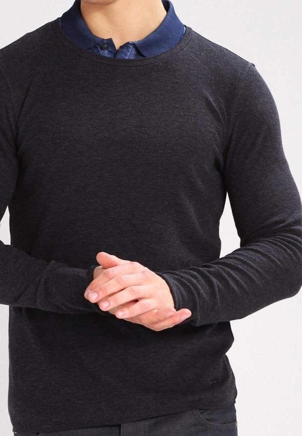 Man som bär en mörkgrå långärmad skjorta över en blå skjorta med krage, händerna lätt hopknäppta vid midjan mot en enkel bakgrund.