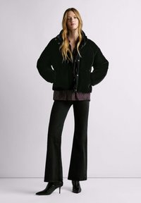 Melna polsterēta jaka ar augstu apkakli, valkāta pār svītrainu blūzi. Kombinēta ar izpletņveida melnām biksēm un melniem, ar papēdi, smailiem zābakiem.