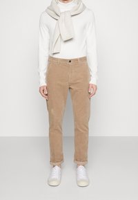 Pantalon en velours côtelé beige à coupe droite, ourlets retroussés, associé à un col roulé blanc et un pull léger porté autour du cou. Baskets blanches.