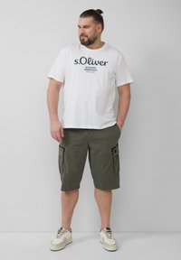 Biała koszulka z okrągłym dekoltem i granatowym nadrukiem; oliwkowe szorty cargo z bocznymi kieszeniami; beżowe sneakersy z czarnymi akcentami.