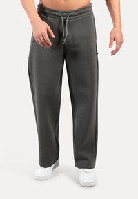 Mies, joka pitää yllä löysiä tummanharmaita jumppahousuja, joissa on nauhakuulolla varustettu vyötärö, ja valkoisia tennarit, seisoo tasasella taustalla.