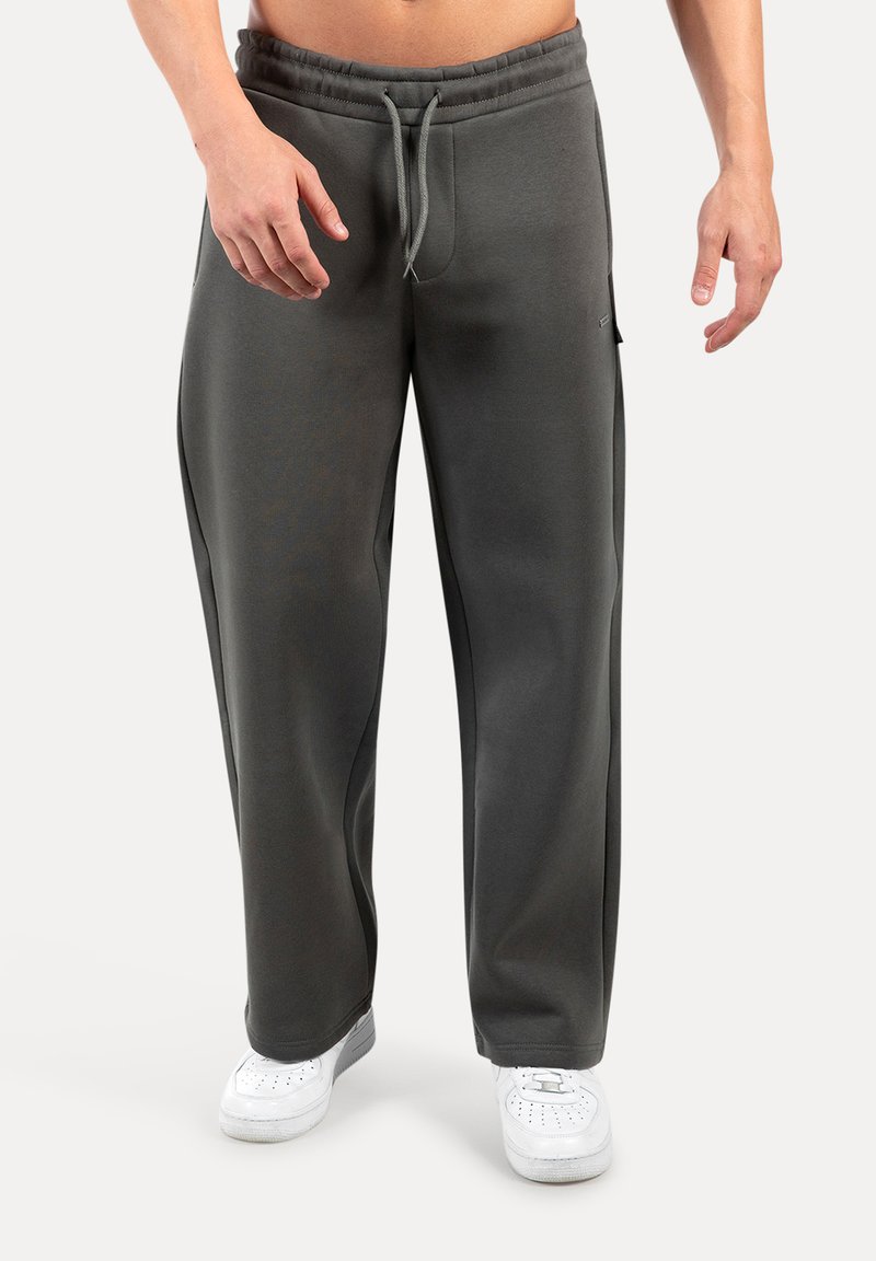 Mies, joka pitää yllä löysiä tummanharmaita jumppahousuja, joissa on nauhakuulolla varustettu vyötärö, ja valkoisia tennarit, seisoo tasasella taustalla.