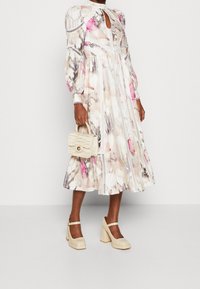 Robe à imprimé floral dans des teintes douces, avec des manches bouffantes, un décolleté en trou de serrure et des accents en dentelle. Associée à un sac à main crème et des talons compensés épais.