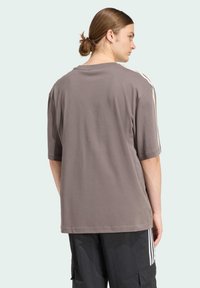 Nadmerné sivé tričko z jemnej látky, s krátkymi rukávmi a jemnými bielymi bočnými pruhmi. Pohľad zozadu s voľným strihom.