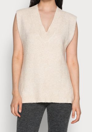 Kobieta w beżowej, bezrękawnikowej, dzianinowej kamizelce z dekoltem w serek, w połączeniu z ciemnoszarymi, teksturowanymi spodniami, stojąca na jednolitym tle.