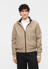 Άνδρας με σγουρά μαλλιά που φοράει μπεζ μπουφάν με φερμουάρ πάνω από λευκό πουκάμισο με λογότυπο EA7 και μαύρο παντελόνι, στέκεται μπροστά σε απλό φωτεινό φόντο.