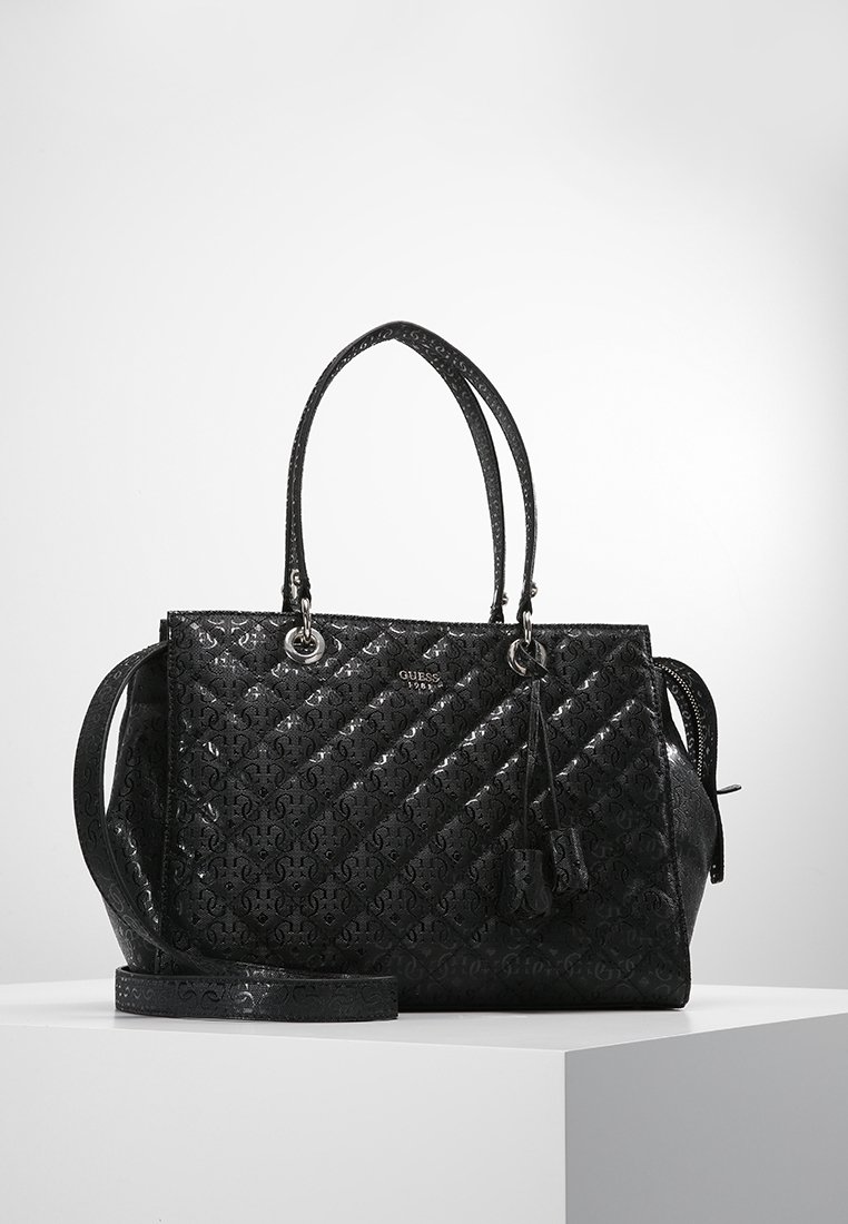 Sac à main en cuir matelassé noir avec double sangle, ferrures argentées, logo Guess et détail de pompon suspendu, posé sur un piédestal blanc.