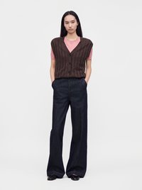 Jeune femme aux longs cheveux foncés portant un gilet sans manches marron boutonné sur une chemise rose, un pantalon large et foncé, et des chaussures noires.
