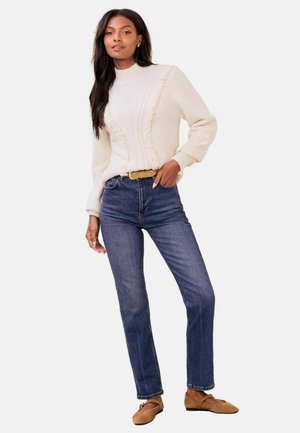 Frau trägt einen cremefarbenen, strukturierten Pullover, dunkelblaue hoch taillierte Jeans, braunen Gürtel und beigefarbene flache Schuhe, steht mit einer Hand in der Tasche.