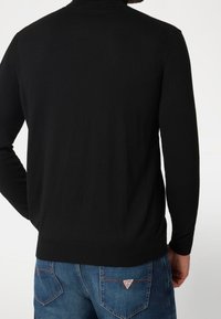 Homme portant un pull noir à manches longues et un jean en denim bleu avec un logo triangulaire Guess sur la poche arrière, de dos.