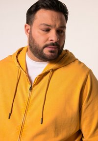 Sudadera amarilla con cremallera y capucha ajustable, fabricada en tejido suave, con una camiseta blanca debajo y herrajes de cremallera minimalistas.