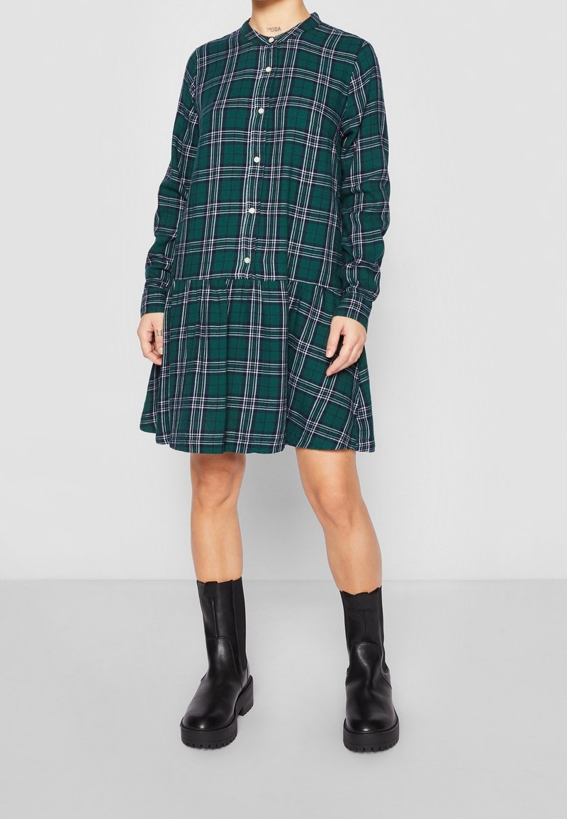 Robe chemise à carreaux verts avec manches longues, boutonnée à l'avant, et jupe à volants. Associée à des bottines noires épaisses, elle présente un col arrondi.
