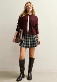 Burgundy cardigan, vit topp och svart rutig kjol, matchat med knähöga bruna läderstövlar och en brun handväska. Släta texturer, lager-på-layer-look.