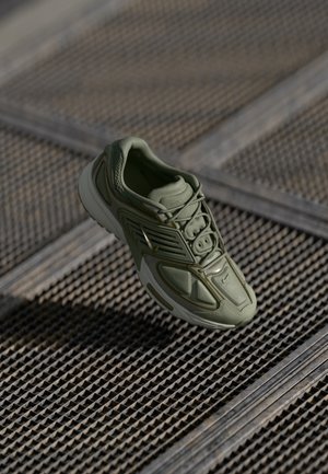 Grøn atletiksneakers med hvid sål, der hviler på en tekstureret metalristflade i blødt naturligt lys.