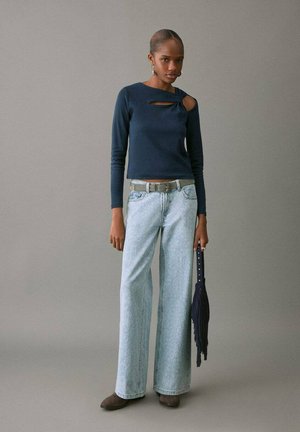 Top blu navy a maniche lunghe con apertura sulle spalle, abbinato a jeans a gamba larga azzurri, accentuato da una cintura grigia e una borsa a frange blu navy.