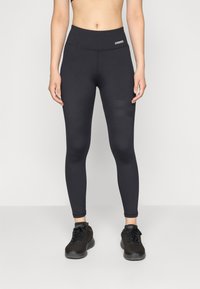 Schwarze Leggings mit hohem Bund, aus elastischem Stoff gefertigt. Besitzt strukturierte Akzente an den Oberschenkeln. Getragen mit schwarzen Sportschuhen.