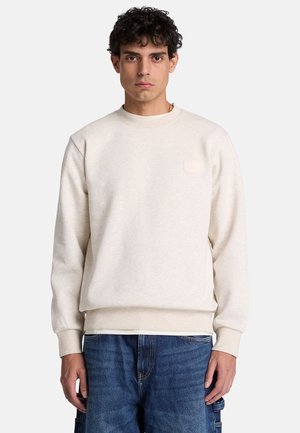 Jonge man met krullend haar, gekleed in een eenvoudige lichtbeige trui en blauwe spijkerbroek, staand tegen een eenvoudige lichte achtergrond.