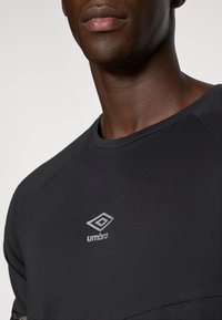 Čierne športové tričko s okrúhlym výstrihom a raglánovými rukávmi, na ľavej hornej strane s bielym logom Umbro. Hladká textúra.