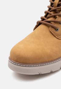 Botine din suede bej, cu vârf rotunjit, șireturi maronii și cusături tonale. Prezintă o talpă din cauciuc gri deschis, cu un finisaj texturat.