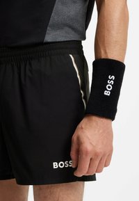 Zwarte sportshorts met wit "BOSS" logo, elastische tailleband en gouden zijrand. Zwarte polsband met witte "BOSS" tekst.