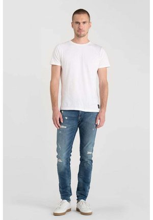 Camiseta blanca de cuello redondo, jeans de mezclilla azul desgastados y zapatillas blancas con suelas beige, modelo de pie contra un fondo claro.