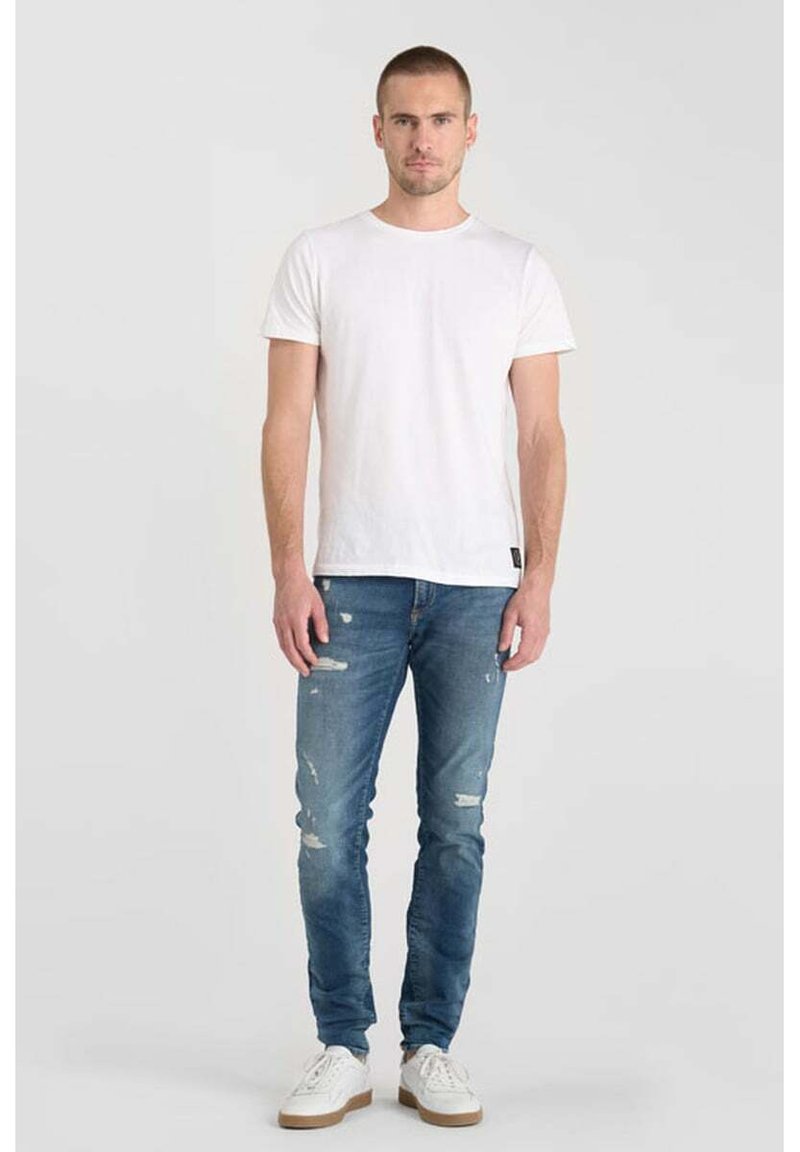 Camiseta blanca de cuello redondo, jeans de mezclilla azul desgastados y zapatillas blancas con suelas beige, modelo de pie contra un fondo claro.