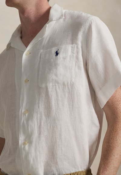 Chemise blanche en lin à manches courtes avec une poche poitrine, ornée d'un logo bleu foncé et de boutons clairs. Texture douce, coupe décontractée.