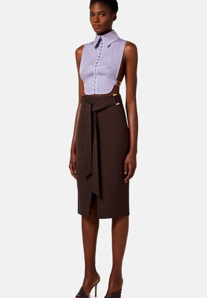 Donna che indossa un top corto senza maniche di colore lilla chiaro con bottoni sul davanti e una gonna midi marrone a vita alta con cintura annodata e spacco, posando con tacchi alti.