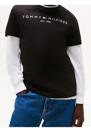 Persona vistiendo una camiseta negra de Tommy Hilfiger sobre una camisa blanca de manga larga y jeans azules, con la mano parcialmente en el bolsillo.