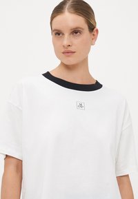 Junge Frau mit zurückgebundenem Haar, die ein weißes T-Shirt mit schwarzem Kragen und kleinem zentralen Logo vor einem einfarbigen Hintergrund trägt.