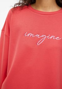 Sudadera color coral de corte holgado, con la palabra "imagine" bordada en cursiva y color rosa. Tejido suave y cuello ancho.
