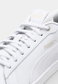 Vit lädersneaker med platta snören, en vadderad krage och vit gummisula. Har präglar "PUMA"-logotypen i guld på tungan och sidan.