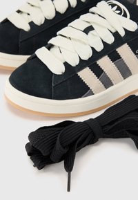 Close-up of dark sneakers with white laces and beige stripes, alongside a bundle of black shoelaces on a light background.