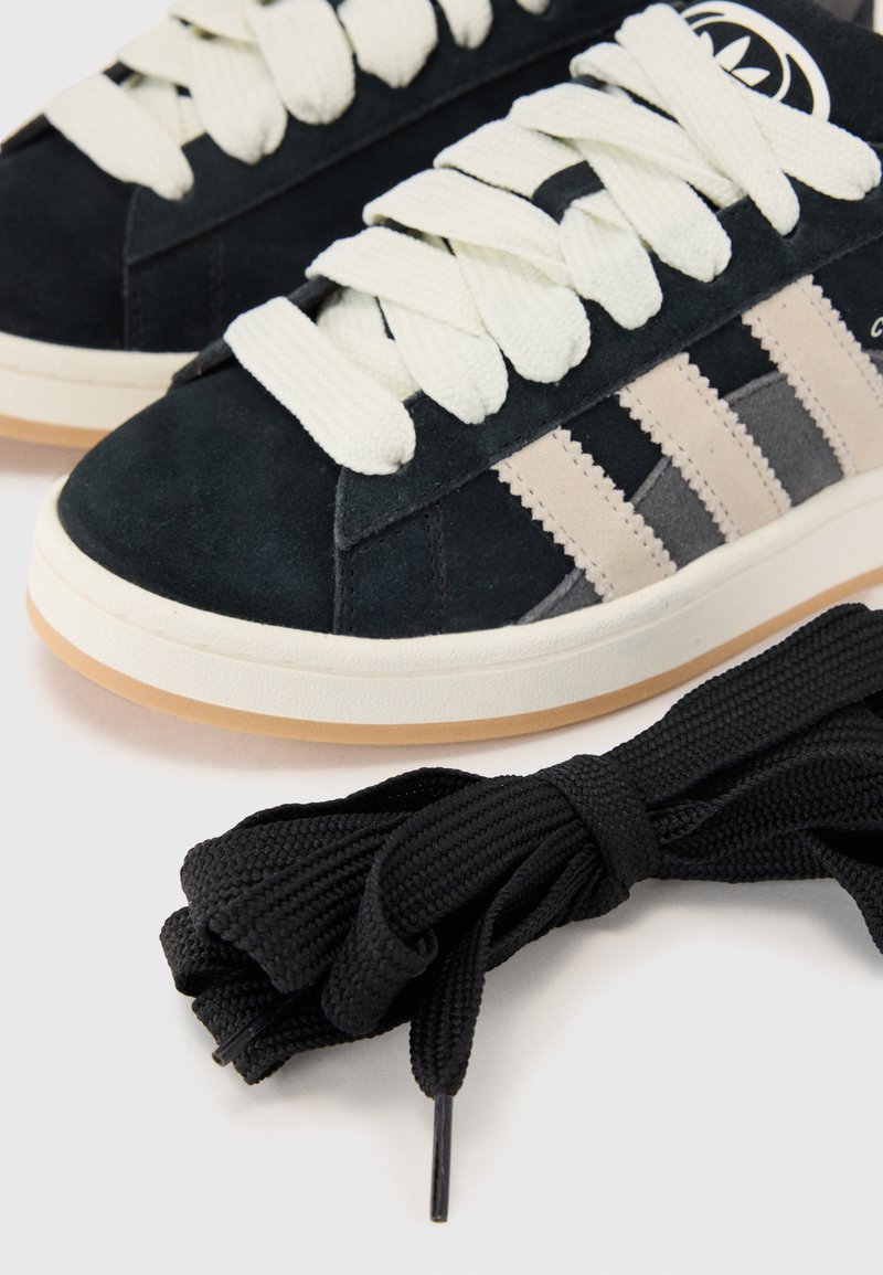 Primer plano de zapatillas oscuras con cordones blancos y rayas beige, junto a un manojo de cordones negros sobre un fondo claro.