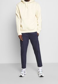 Un sweat à capuche jaune clair avec une poche kangourou, associé à un pantalon tapered marine. Des baskets blanches avec des accents de violet et de noir.