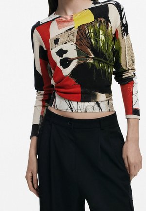 Mujer con una blusa de manga larga con estampado abstracto multicolor combinada con pantalones negros de talle alto, manos descansando cerca de las caderas.