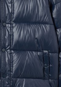 Giacca imbottita blu navy con texture lucida e trapuntata; presenta bottoni a scatto blu e una tasca laterale. Design compatto e isolato per calore.