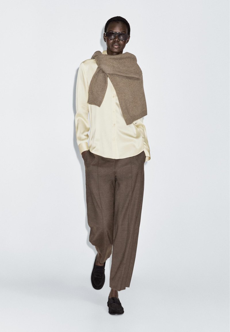 Hnědý vlněný šál přehozený přes krémovou košili s knoflíky, doplněný hnědými kalhotami s vysokým pasem a černými mokasíny. Hladké textury, neutrální barvy.