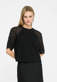 Top nero con un corpo in tessuto solido e maniche in pizzo ricamato trasparente. Le maniche hanno un bordo a scallop ed un motivo floreale.