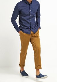 Homme marchant, les mains dans les poches, portant une chemise boutonnée bleu marine à motifs, un pantalon beige retroussé et des chaussures bleu à enfiler.