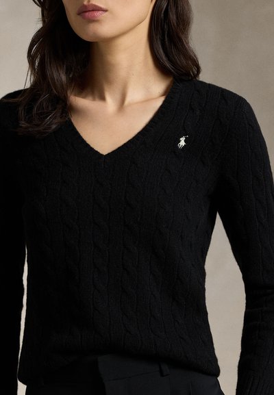 Vrouw draagt een zwarte kabelgebreide V-hals trui met een klein wit geborduurd logo op de linkerborst.