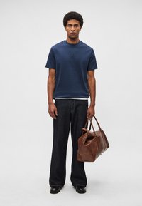 Tumši zils īsroku T-krekls, tumši džinsi un brūna ādas ceļojuma soma. Somai ir divas rokturi un gluda virsma.