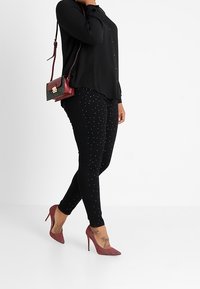 Zwarte blouse met knopen, gecombineerd met zwarte spijkerbroek met zilveren studaccenten. Draag het met rode studded hoge hakken en een kleine rode crossbody tas.