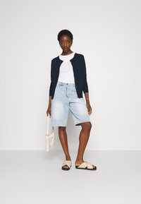 Marine cardigan, witte t-shirt, lichtblauwe denimshorts en beige slip-on sandalen. Houdt een crèmekleurige, geweven handtas vast. Eenvoudige, casual outfit.