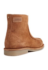 Braune Wildleder-Stiefelette mit abgerundeter Zehenpartie, minimaler Naht und einem Markenpatch auf der Rückseite. Flache Gummisohle mit glatter Oberfläche.