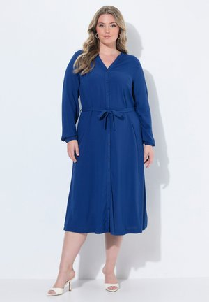 Femme aux longs cheveux blonds portant une robe midi bleue boutonnée et ceinturée ainsi que des talons ouverts blancs, debout devant un fond blanc.
